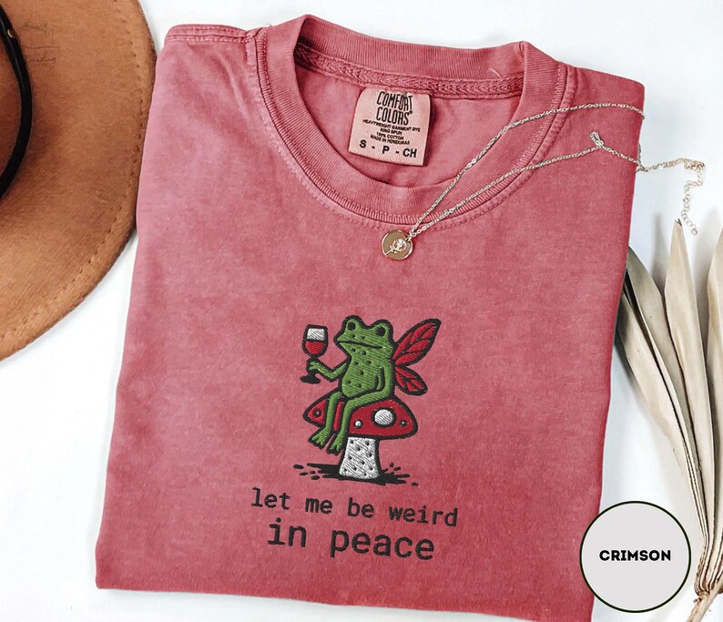 May include: Crimson-colored t-shirt with a crew neck. The shirt features an embroidered design of a green frog with fairy wings, holding a wine glass, sitting on a mushroom. The text "let me be weird in peace" is below the frog.