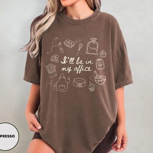 May include: Espresso-colored t-shirt with the text "I'll be in my office" and baking-related illustrations. The design includes a mixer, whisk, oven mitt, apron, and other baking tools. The shirt is a comfortable, oversized fit.