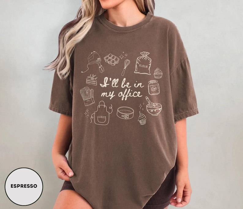May include: Espresso-colored t-shirt with the text "I'll be in my office" and baking-related illustrations. The design includes a mixer, whisk, oven mitt, apron, and other baking tools. The shirt is a comfortable, oversized fit.
