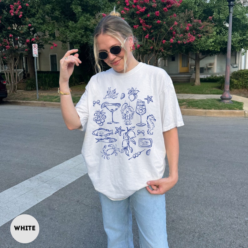 May include: White t-shirt with a blue nautical-themed graphic. The design includes illustrations of seafood, cocktails, and other coastal elements. The shirt is a relaxed fit.