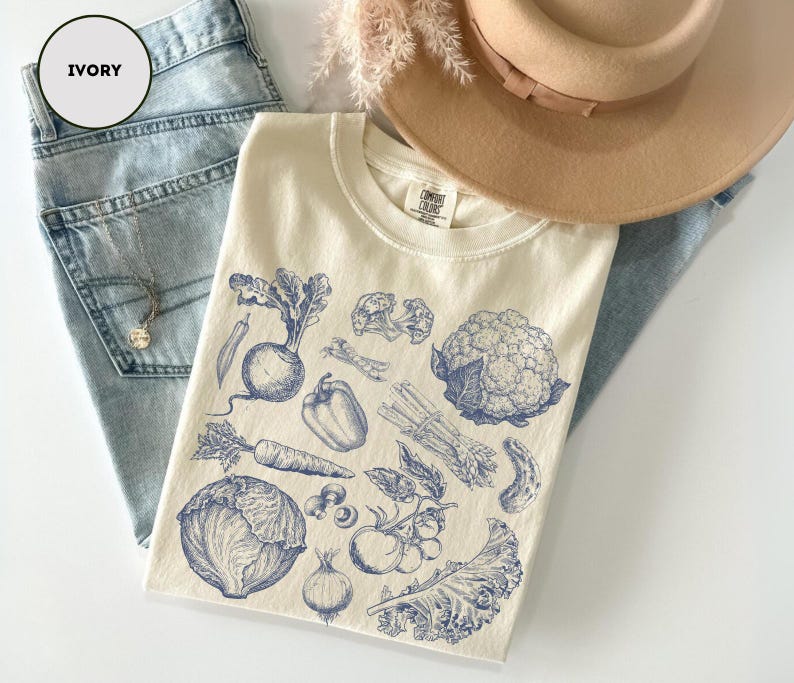 May include: Ivory-colored t-shirt with a blue illustration of various vegetables, including a carrot, cauliflower, and cabbage. The shirt is laid on top of light blue jeans and a tan hat. The shirt has the text "COMFORT COLORS" on the tag.