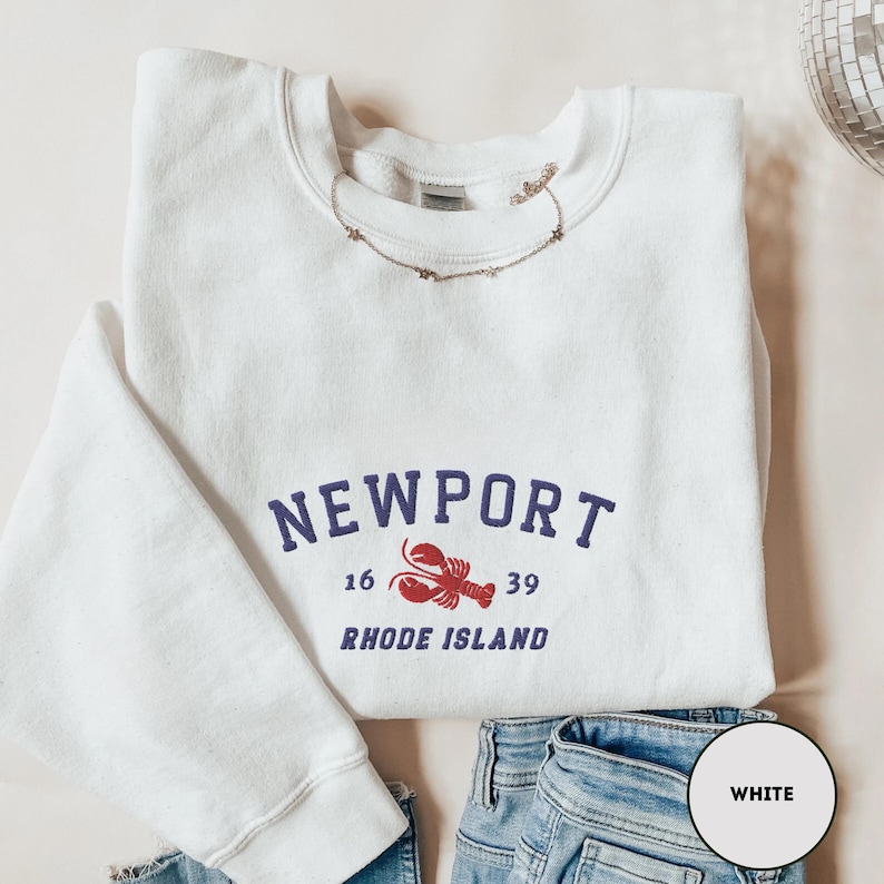 May include: White crew neck sweatshirt featuring the word "NEWPORT" in navy blue, a red lobster graphic, and the text "RHODE ISLAND." The sweatshirt is folded with blue jeans.