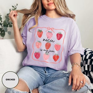 May include: A light purple t-shirt with a strawberry graphic and the text "nicu is my jam". The graphic features nine strawberries in various shades of red and pink, framed by a pink border.
