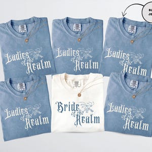 May include: A collection of blue jean t-shirts and one cream-colored t-shirt. The blue shirts have the text "Ladies of the Realm" printed in white. The cream shirt says "Bride of the Realm" in blue. Each shirt has a gold necklace.
