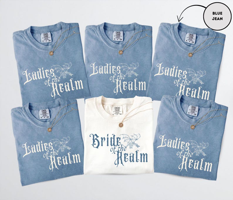 May include: A collection of blue jean t-shirts and one cream-colored t-shirt. The blue shirts have the text "Ladies of the Realm" printed in white. The cream shirt says "Bride of the Realm" in blue. Each shirt has a gold necklace.