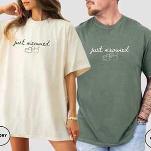 May include: Ivory and moss green t-shirts with the text "just meowied" and a cat illustration. The ivory shirt is worn by a person with a gold bracelet. The moss green shirt is worn by a person with tattoos on their arm.