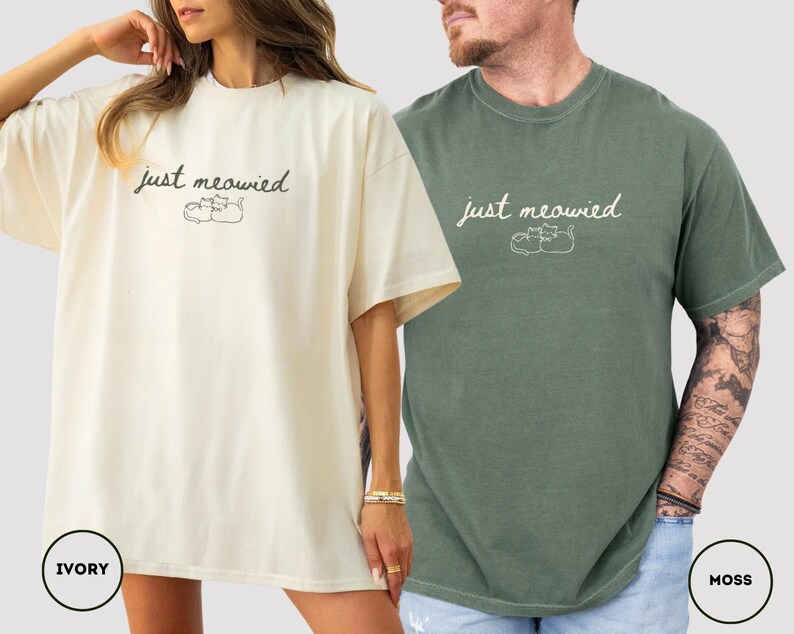 May include: Ivory and moss green t-shirts with the text "just meowied" and a cat illustration. The ivory shirt is worn by a person with a gold bracelet. The moss green shirt is worn by a person with tattoos on their arm.