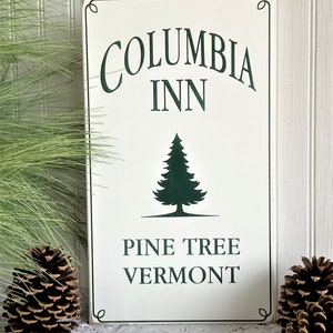 May include: A rectangular white wooden sign with dark green text and a pine tree graphic. The text reads COLUMBIA INN, PINE TREE, and VERMONT. The sign features a thin dark green border with decorative corner accents and a distressed finish.