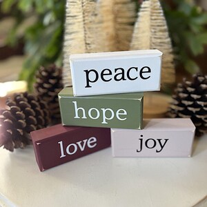 May include: Four rectangular wooden blocks stacked together. The top white block reads peace, the middle olive green block reads hope, the bottom left maroon block reads love, and the bottom right light pink block reads joy in black and white text.