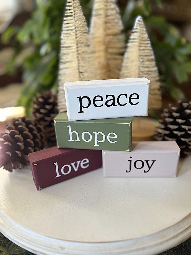 May include: Four rectangular wooden blocks stacked together. The top white block reads peace, the middle olive green block reads hope, the bottom left maroon block reads love, and the bottom right light pink block reads joy in black and white text.