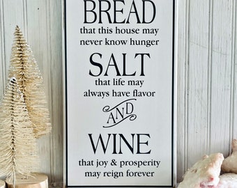 Könnte beinhalten: Ein weißes Holzschild mit schwarzem Text, der lautet: "BREAD that this house may never know hunger SALT that life may always have flavor AND WINE that joy & prosperity may reign forever"