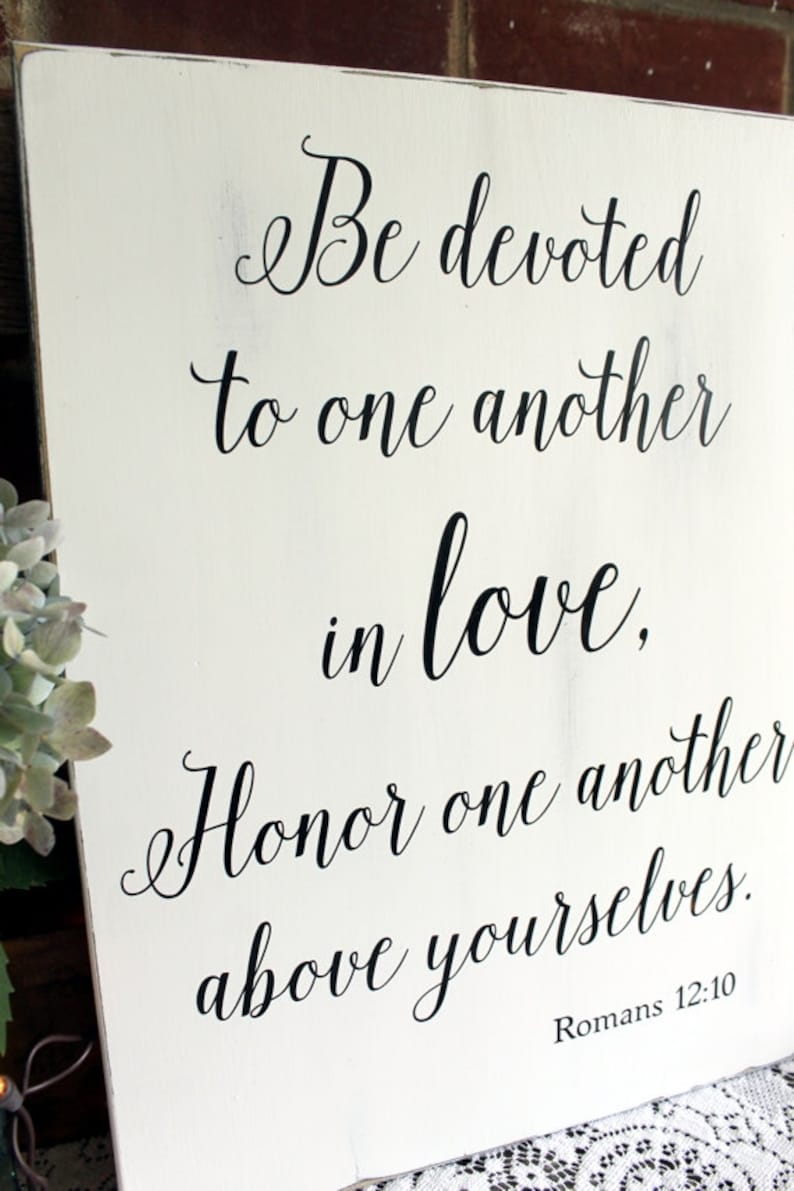 May include: White wooden sign with black calligraphy text that reads "Be devoted to one another in love, Honor one another above yourselves. Romans 12:10".