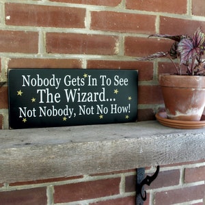 Black wood sign with white lettering saying, Nobody Gets In To See The Wizard...Not Nobody, Not No How. Gold stars. 6x14 inches.