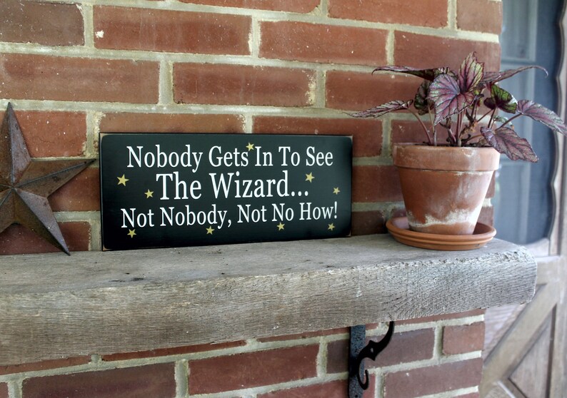 Black wood sign with white lettering saying, Nobody Gets In To See The Wizard...Not Nobody, Not No How. Gold stars. 6x14 inches.
