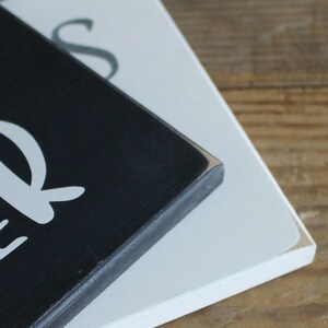 May include: Close-up of a black wooden sign with white lettering that reads "Our" and "Bless" in a cursive font. The sign is stacked on top of a white wooden sign.