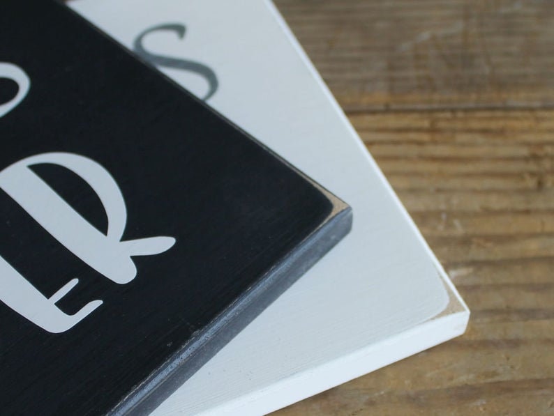 May include: Close-up of a black wooden sign with white lettering that reads "Our" and "Bless" in a cursive font. The sign is stacked on top of a white wooden sign.