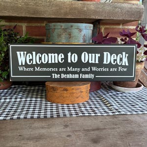 May include: A rectangular black sign with white text that reads "Welcome to Our Deck Where Memories are Many and Worries are Few - The Denham Family." The sign is displayed on a wooden surface.