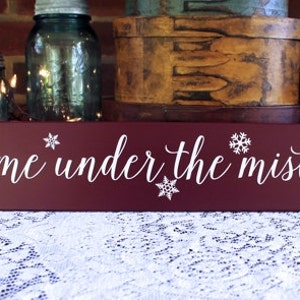 May include: A rectangular, burgundy wooden sign with the phrase "meet me under the mistletoe" in white script. White snowflake designs are placed around the text. The sign is a festive holiday decoration.