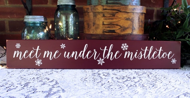 May include: A rectangular, burgundy wooden sign with the phrase "meet me under the mistletoe" in white script. White snowflake designs are placed around the text. The sign is a festive holiday decoration.