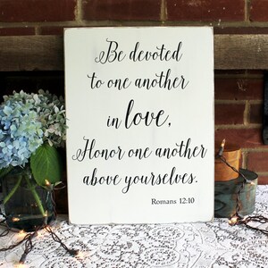 May include: White wooden sign with black calligraphy text that reads "Be devoted to one another in love, Honor one another above yourselves. Romans 12:10".