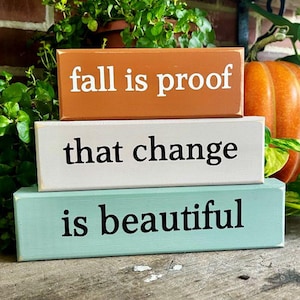 May include: Three rectangular wooden blocks stacked vertically. The top block is burnt orange with white text reading fall is proof. The middle block is off-white with black text reading that change. The bottom block is light teal with black text reading is beautiful.