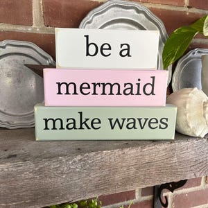May include: Three rectangular wooden blocks stacked on a wooden shelf. The top block is white with the words "be a" in black. The middle block is pink with "mermaid" in black. The bottom block is light green with "make waves" in black.