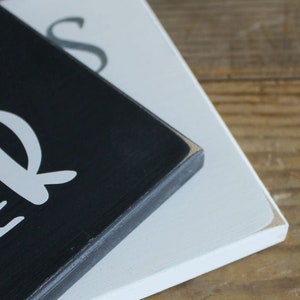May include: Close-up of a black wooden sign with white lettering that reads "Our" and "Bless" in a cursive font. The sign is stacked on top of a white wooden sign.