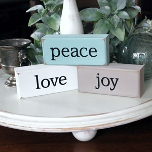 May include: Three wooden blocks stacked on a white tiered tray. The top block is light blue and says "peace" in black lettering. The middle block is white and says "love" in black lettering. The bottom block is light brown and says "joy" in black lettering.