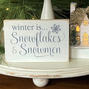 May include: White wooden sign with the text "winter is... snowflakes & snowmen" in gray lettering. A snowflake graphic is above the text. The sign is sitting on a white tiered tray.