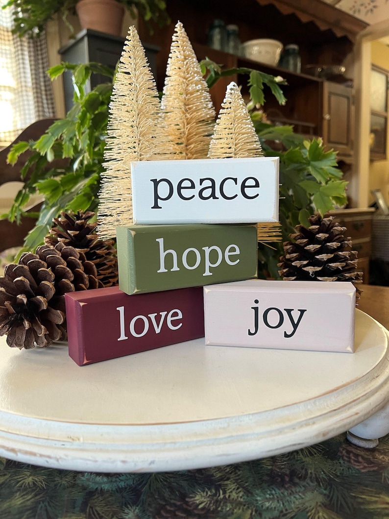May include: A set of four rectangular wooden blocks stacked together. The top block is white with the word peace, the second is olive green with hope, the third is maroon with love, and the fourth is light pink with the word joy in black.
