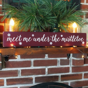 May include: A rectangular, burgundy wooden sign with the phrase "meet me under the mistletoe" in white script. The sign is decorated with white snowflake accents. The sign is displayed on a wooden shelf with Christmas decorations.