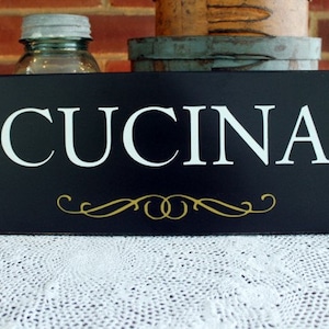 Puede incluir: Un letrero rectangular negro con la palabra "CUCINA" en letras may&uacute;sculas blancas. Debajo del texto hay un adorno dorado. El letrero est&aacute; sobre un mantel de encaje blanco.