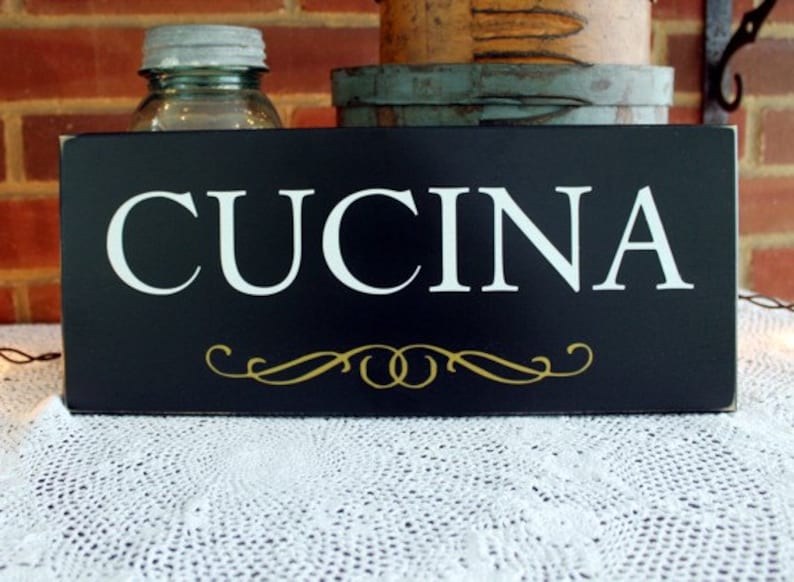 Puede incluir: Un letrero rectangular negro con la palabra "CUCINA" en letras may&uacute;sculas blancas. Debajo del texto hay un adorno dorado. El letrero est&aacute; sobre un mantel de encaje blanco.