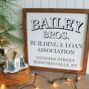 May include: A square wooden sign with a dark brown frame and a white background featuring black text that reads BAILEY BROS. BUILDING & LOAN ASSOCIATION GENESEE STREET BEDFORD FALLS, NY in a mix of bold serif and sans-serif fonts.