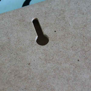 May include: A close-up of a light brown wooden surface with a single, vertical, rectangular hole cut out of it. The hole is centered in the image and is slightly wider at the top than at the bottom.