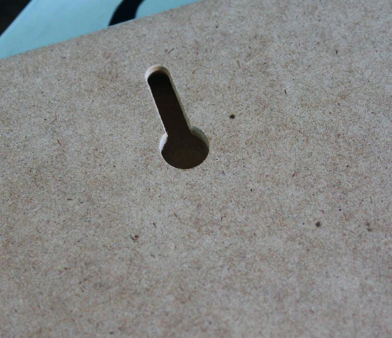 May include: A close-up of a light brown wooden surface with a single, vertical, rectangular hole cut out of it. The hole is centered in the image and is slightly wider at the top than at the bottom.