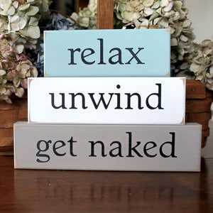 May include: Three rectangular wooden signs stacked on top of each other. The top sign is light blue with the word "relax" in black. The middle sign is white with "unwind" in black. The bottom sign is gray with "get naked" in black.