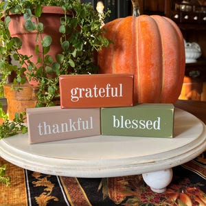 May include: A fall-themed display featuring three wooden blocks with the words "grateful," "thankful," and "blessed" in white lettering. An orange pumpkin and a terracotta pot with greenery are also present.