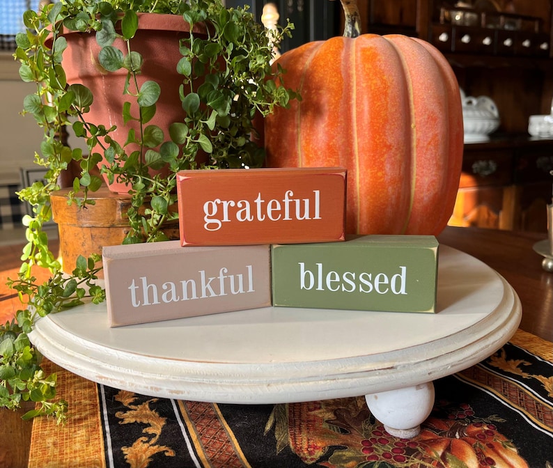 May include: A seasonal arrangement with three wooden blocks inscribed with "grateful," "thankful," and "blessed." An orange pumpkin and a terracotta pot with greenery are also present, creating a warm autumn scene.
