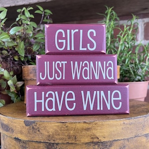 May include: Three stacked, rectangular wooden blocks with white text on a burgundy background. The top block reads "GIRLS", the middle block says "JUST WANNA", and the bottom block says "HAVE WINE".