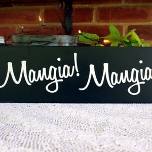 K&ouml;nnte beinhalten: Schwarzes Holzschild mit wei&szlig;er Aufschrift "Mangia! Mangia!" Das Schild steht auf einer wei&szlig;en Spitzentischdecke.