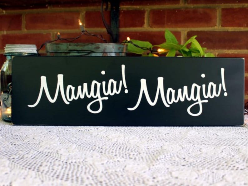 K&ouml;nnte beinhalten: Schwarzes Holzschild mit wei&szlig;er Aufschrift "Mangia! Mangia!" Das Schild steht auf einer wei&szlig;en Spitzentischdecke.