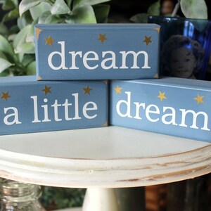 May include: Three blue wooden blocks with gold stars and white lettering. The blocks spell out "dream a little dream".