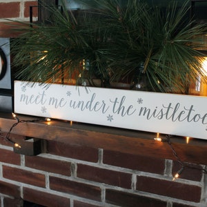 May include: A white rectangular wooden sign with the phrase "meet me under the mistletoe" in gray script. The sign is decorated with snowflakes and sits on a wooden mantel, with Christmas lights and greenery in the background.