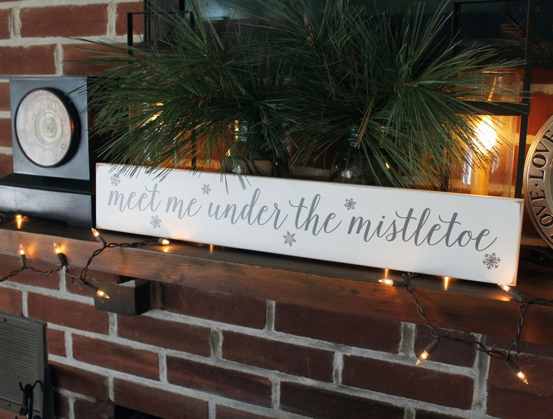 May include: A white rectangular wooden sign with the phrase "meet me under the mistletoe" in gray script. The sign is decorated with snowflakes and sits on a wooden mantel, with Christmas lights and greenery in the background.