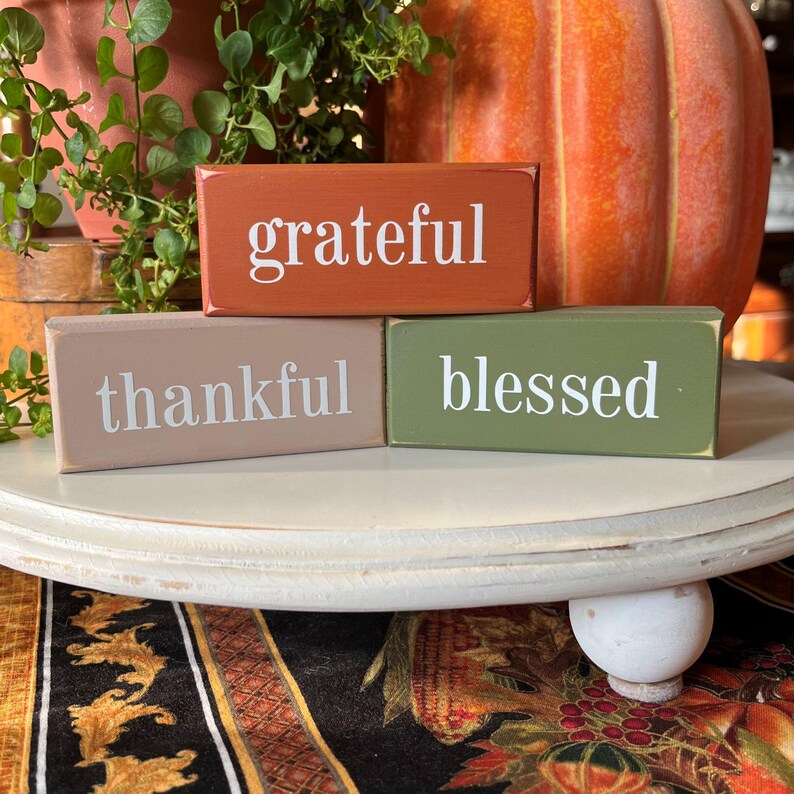 May include: Three rectangular wooden blocks with the words "grateful", "thankful", and "blessed" in white lettering. The blocks are painted in shades of orange, tan, and green. A large orange pumpkin and greenery are in the background.