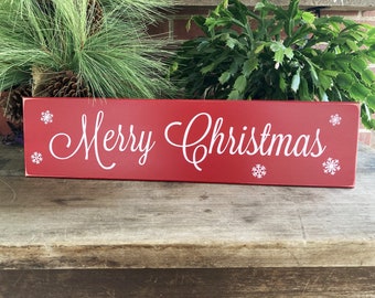 May include: A rectangular red wooden sign with the words "Merry Christmas" in white script. White snowflake designs are placed around the text. The sign is a festive holiday decoration.