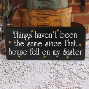 May include: Black wooden sign with gold stars and white text that reads "Things haven't been the same since that house fell on my sister."