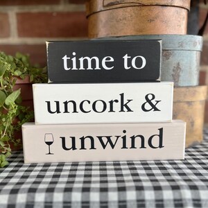 May include: Three wooden signs stacked on top of each other. The top sign is black with white text that reads "time to". The middle sign is white with black text that reads "uncork &". The bottom sign is light brown with black text that reads "unwind" and a small wine glass icon.
