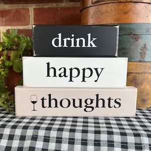 May include: Three wooden blocks stacked on top of each other. The top block is black with white text that says "drink". The middle block is white with black text that says "happy". The bottom block is light pink with black text that says "thoughts" and a black wine glass icon.